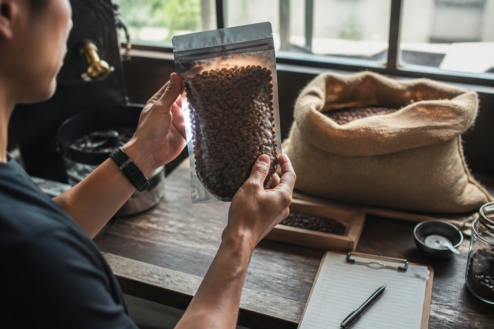 A person holding a bag of coffee beans up to the light, examining the roast date on the label, with a burlap sack of beans in the background