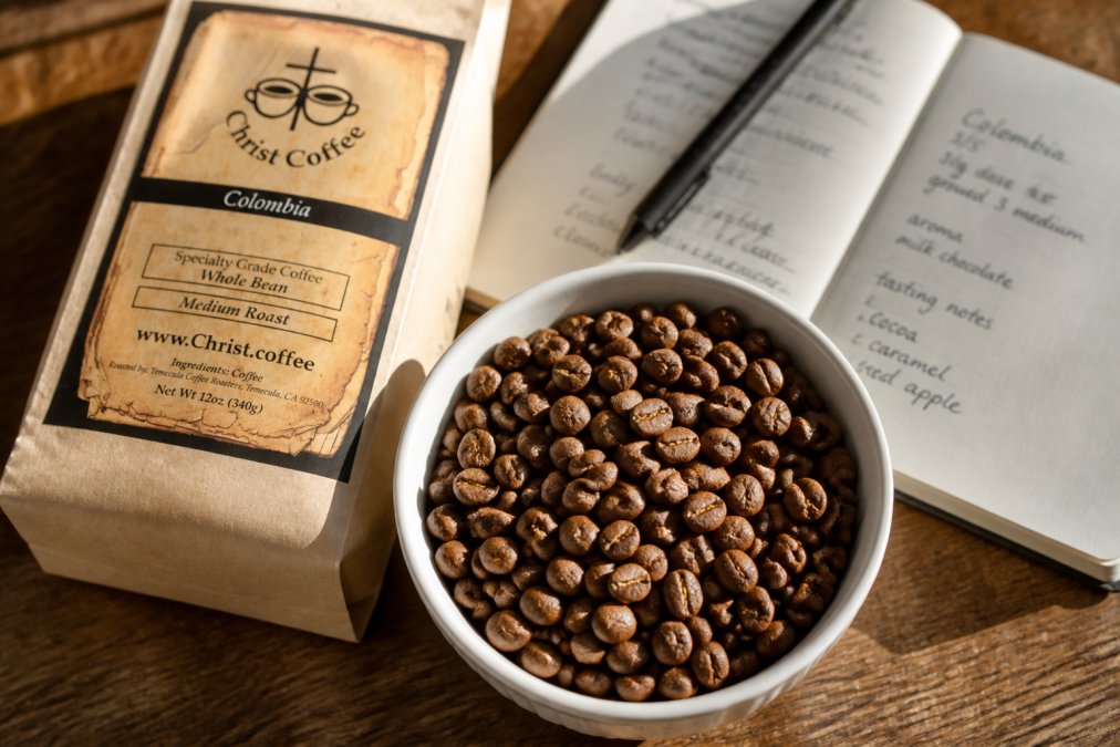 Close-up of freshly roasted specialty coffee beans in a white ceramic cupping bowl, with individual beans showing varied brown tones and texture, alongside an open notebook with handwritten tasting notes and a wooden table with natural afternoon light streaming across