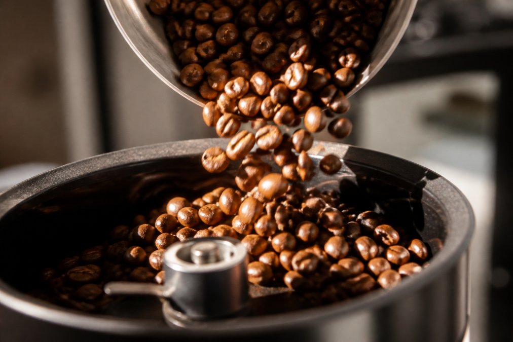 Close-up of specialty coffee beans cascading into a grinder, showing rich brown and amber tones with morning sunlight streaming across the beans