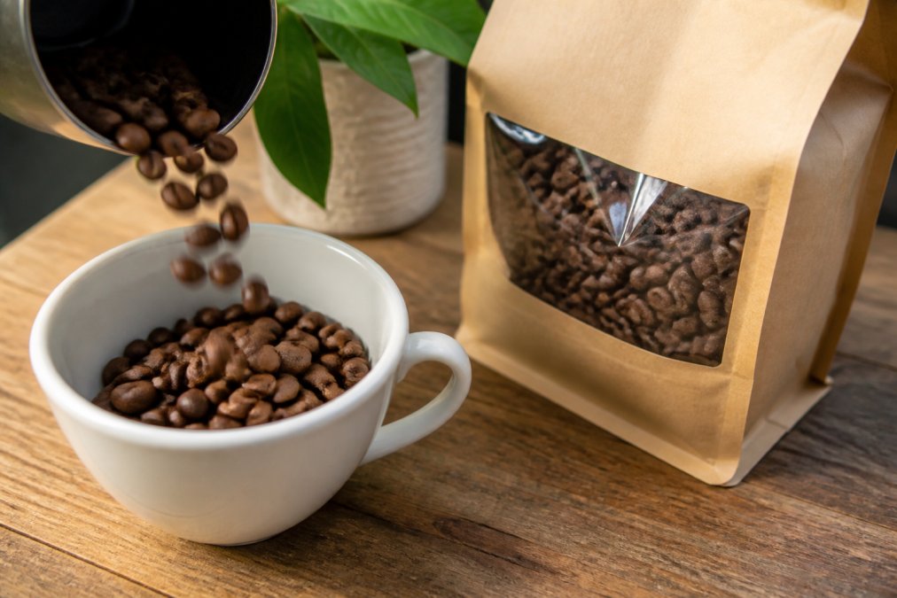 Close-up of specialty coffee beans being poured into a white ceramic cup, showing rich brown color and texture, with a roasted coffee bag displaying origin information and roast date visible in soft natural light