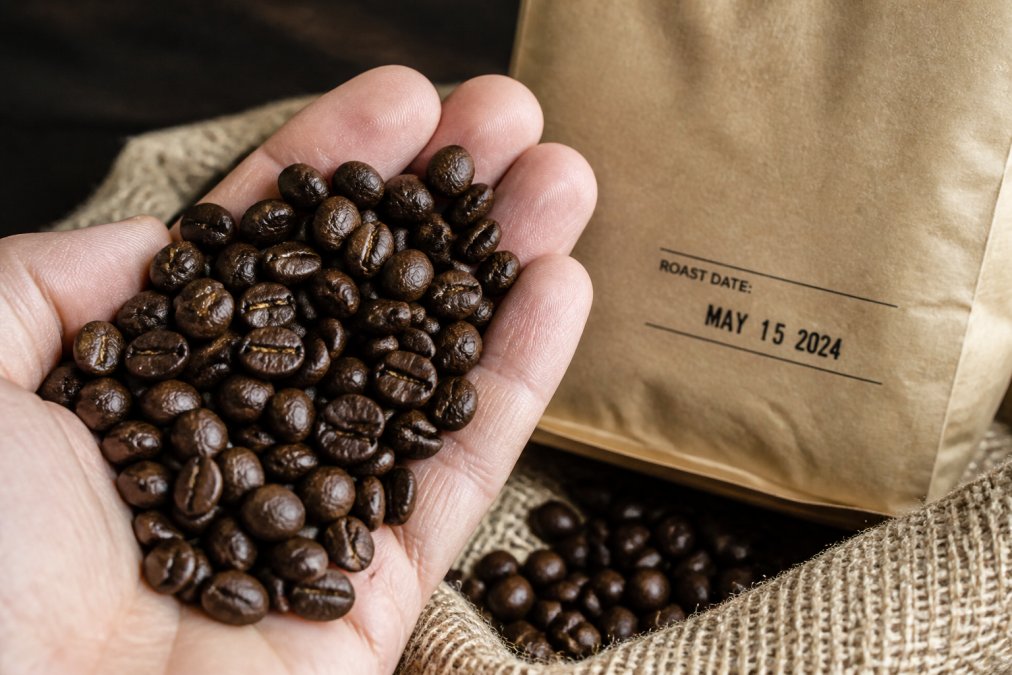 Close-up of freshly roasted specialty coffee beans in a hand, showing glossy surface and rich dark color, with roast date label visible on the bag in background