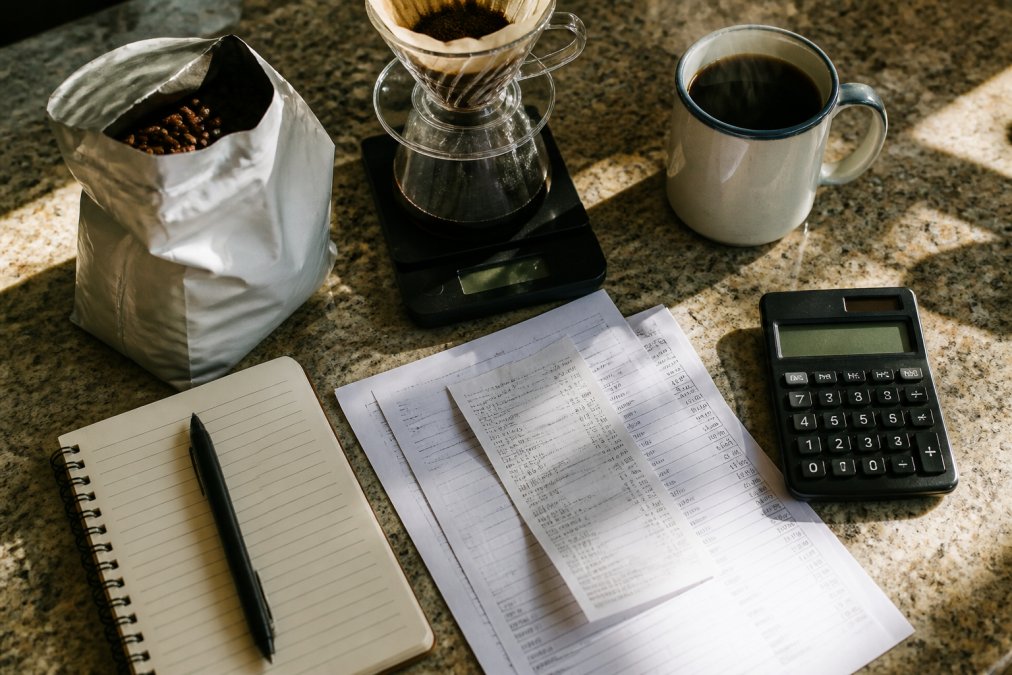 Overhead shot of a kitchen counter with a digital scale, a bag of whole bean coffee, a pour-over dripper, and a steaming mug, natural morning light