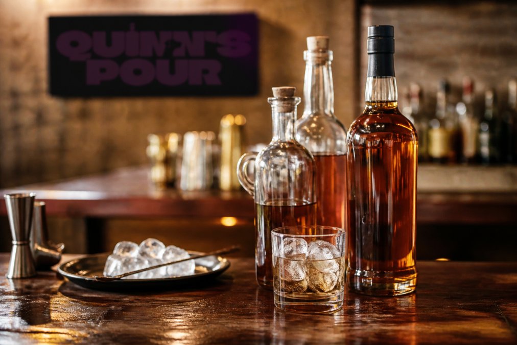 Close-up of Quinn's Pour spiced rum bottles arranged on a wooden bar counter with ice and a glass, warm ambient lighting