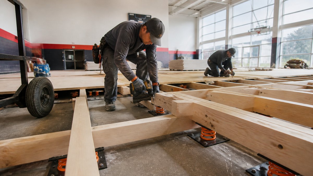 Professional installers positioning power springs and fastening systems on a concrete base in a gymnasium, with visible subfloor structure and installation equipment in natural daylight