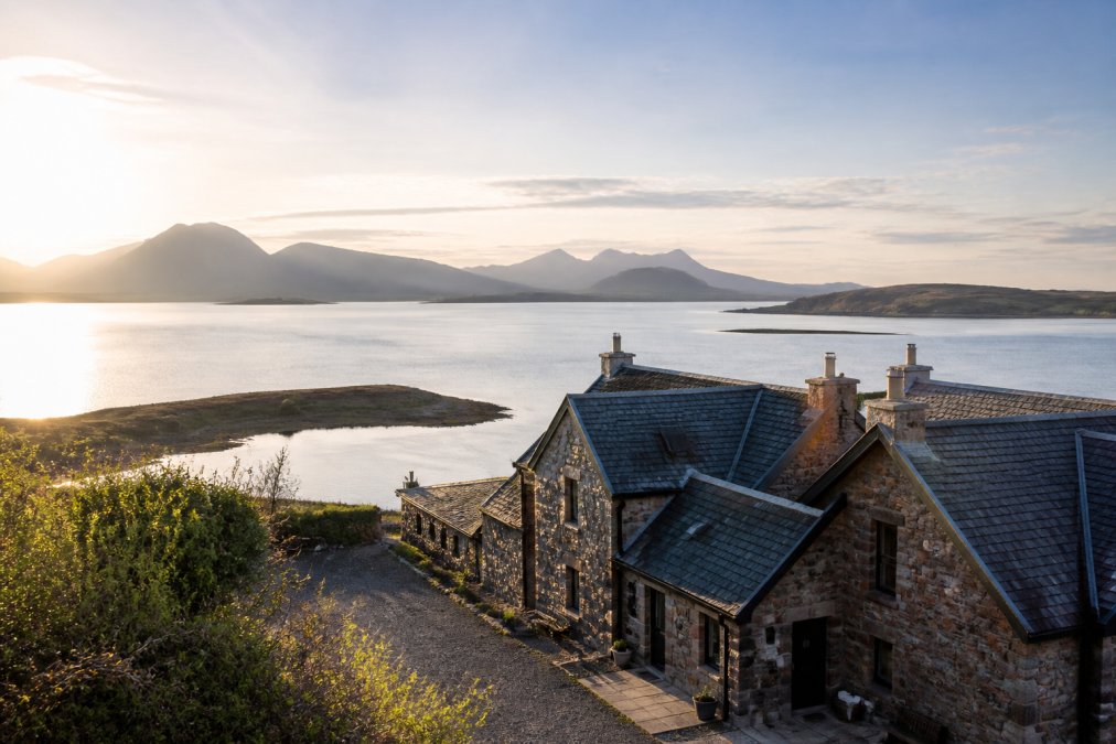 Westerly coastal landscape of the Isle of Skye with stone lodge buildings overlooking a loch, morning light illuminating the water and surrounding mountains in the distance