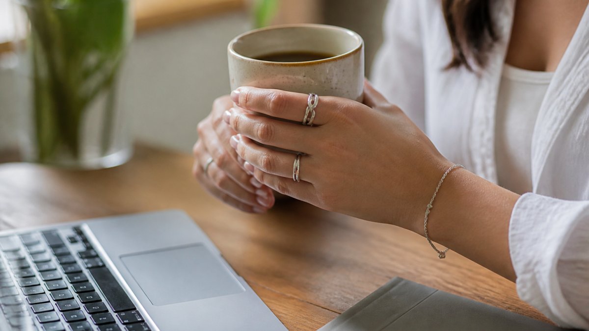 Close-up of hands wearing sterling silver rings and a delicate bracelet while holding a warm coffee cup at a desk, with a laptop keyboard visible in soft natural window lighting