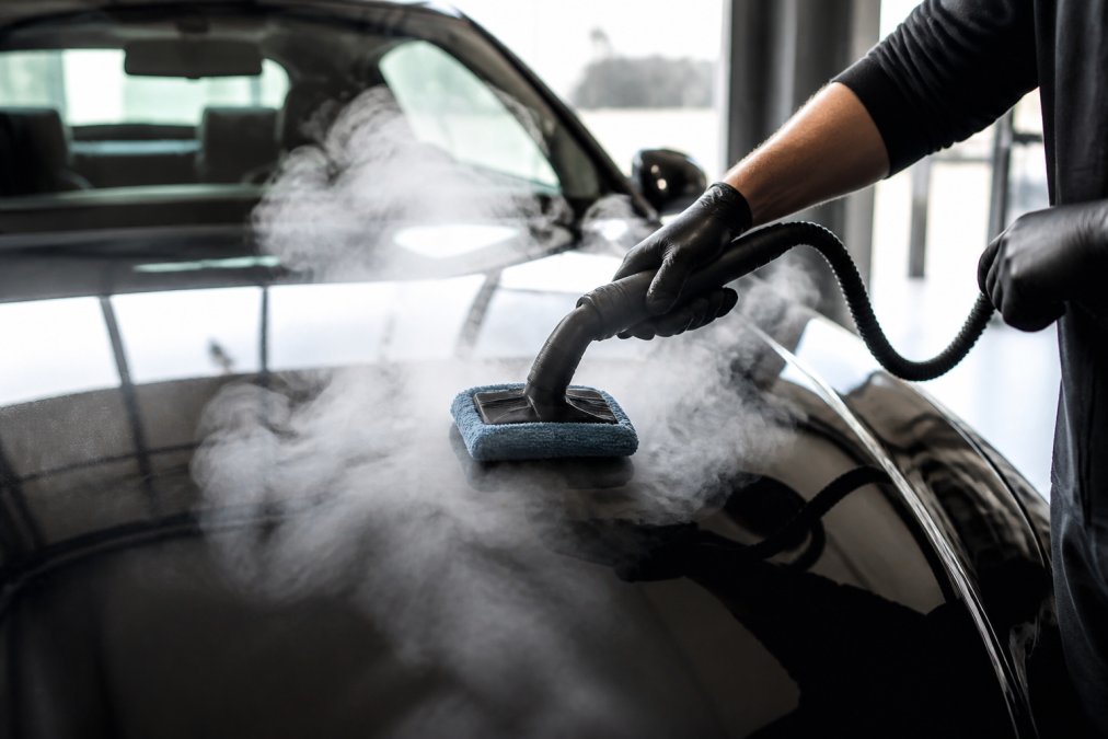 Close-up van iemand die een stoomreiniger met microvezeldoek over een glanzende motorkap beweegt, zichtbare stoomwolk, garage met natuurlijk licht
