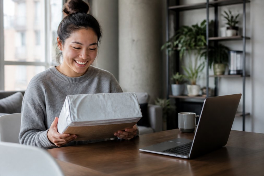 A young woman in a bright Canadian apartment holding a cardboard parcel wrapped in plastic shipping mailer, with a laptop showing a tracking page on the table beside her