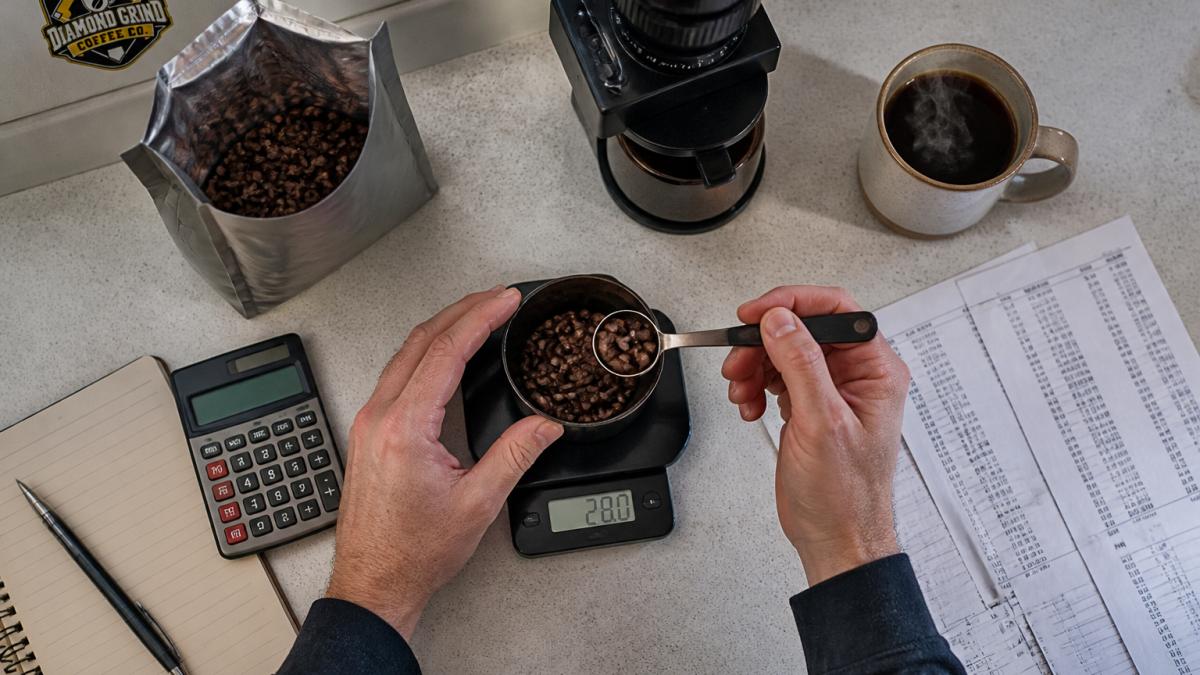 Hands measuring coffee beans on a digital scale next to a grinder and a steaming mug of fresh coffee