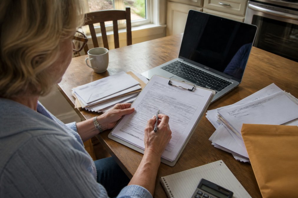 A person at a kitchen table reviewing insurance paperwork with a laptop open, highlighting a claim form with a pen, soft natural light from a window