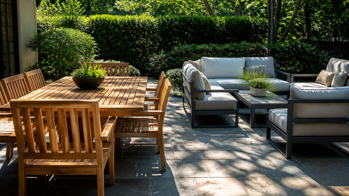 A sunlit patio with a teak dining table and chairs beside a sleek aluminum lounge set, cushions in place, dappled afternoon light through nearby trees