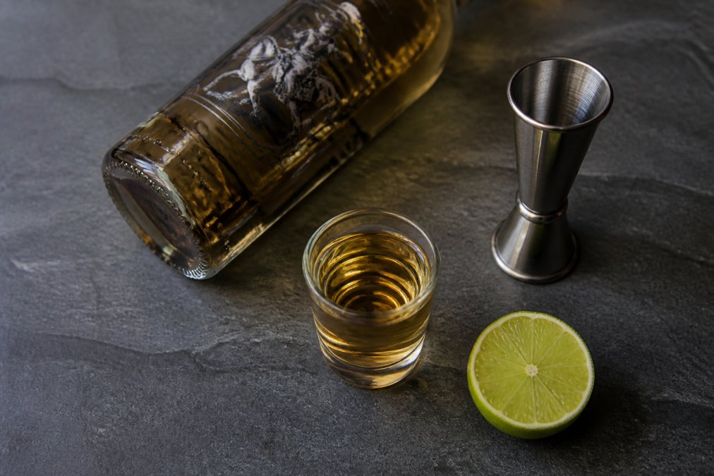 Overhead shot of a 25ml shot glass filled with golden tequila alongside a Thiago tequila bottle on a dark slate surface, with a small stainless steel jigger and a halved lime resting nearby, studio lighting casting soft shadows