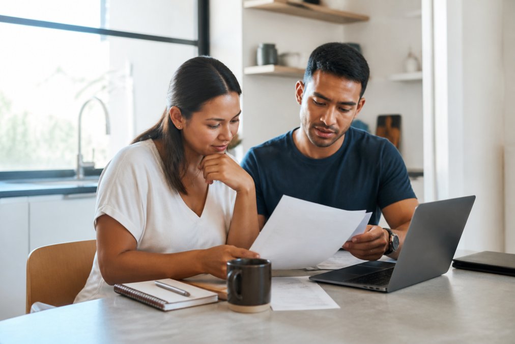 A young professional couple reviewing financial documents on a laptop at a bright, modern kitchen table, with a coffee mug and notebook nearby