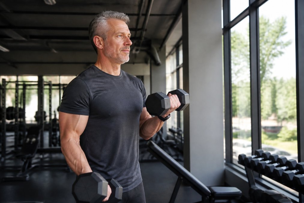 A fit man in his early 50s performing dumbbell curls in a well-lit gym, looking focused and energetic, with natural light coming through large windows