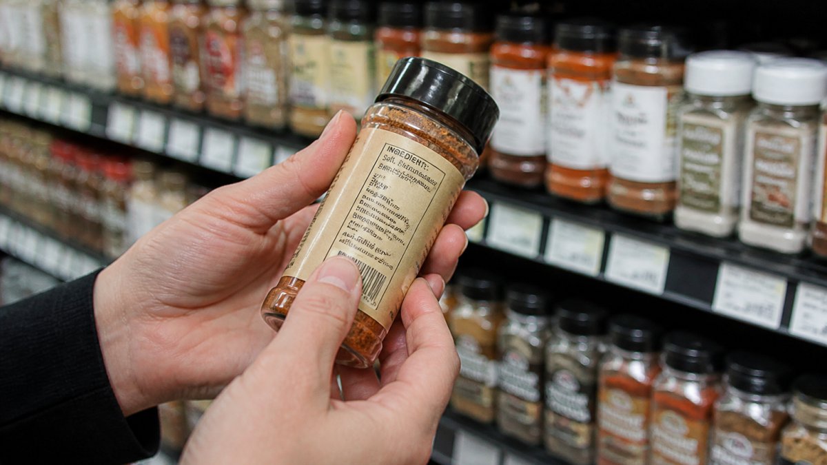 Person in a grocery store aisle reading the ingredient label on a glass spice jar