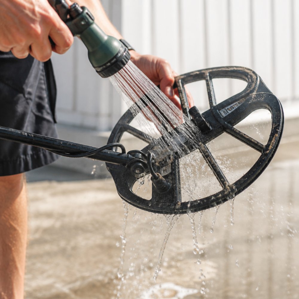 Close-up of hands rinsing metal detector coil with fresh water from hose, showing water streaming over coil surface and sand particles washing away