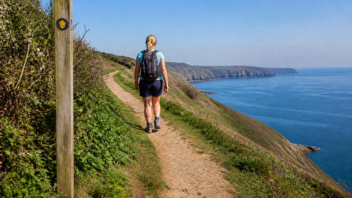 A beginner walker on a coastal path with gentle terrain, clear waymarking visible, and dramatic sea cliffs visible in the distance on a bright, clear day