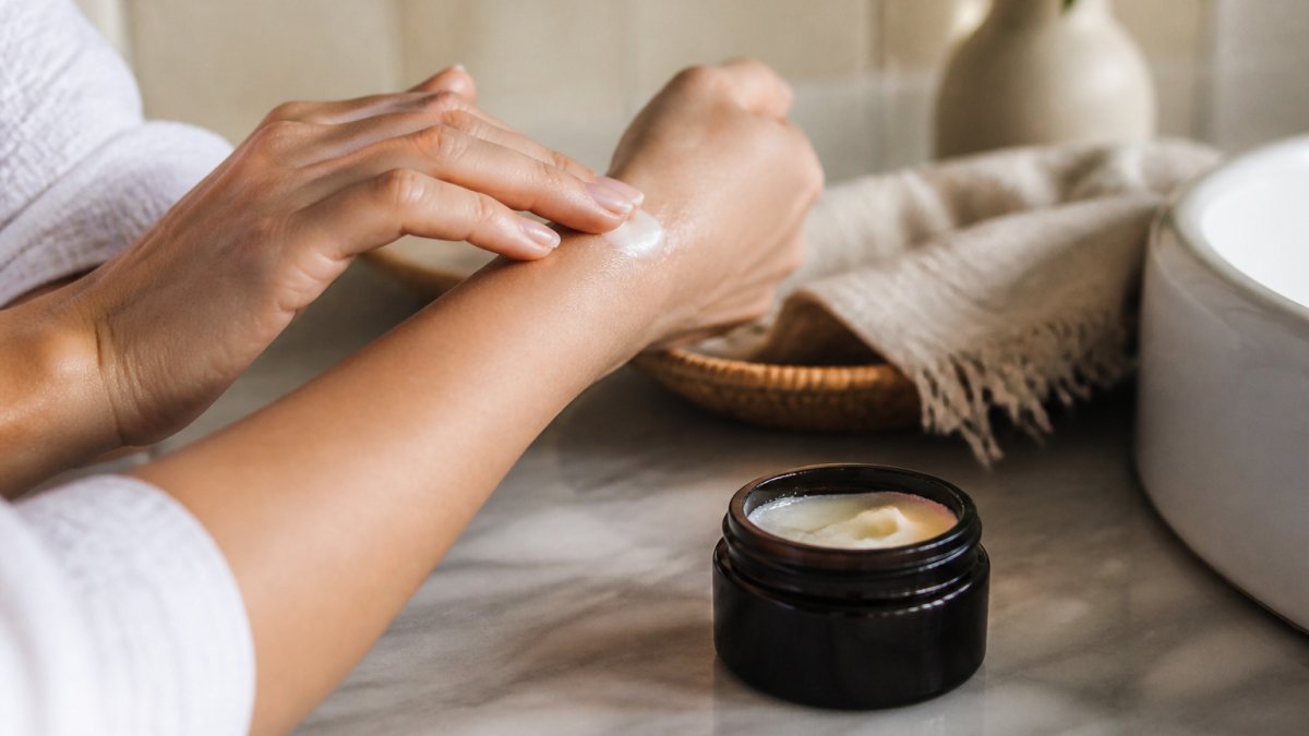 Close-up of hands applying a small amount of creamy white tallow balm to a forearm, soft natural light, clean bathroom counter setting