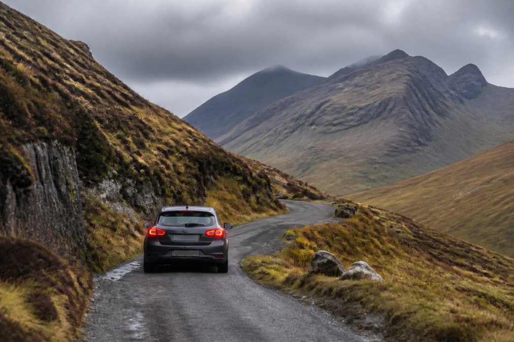 Winding single-track Highland road cutting through dramatic mountain scenery with a car navigating carefully around a bend, passing places visible ahead, cloudy sky overhead