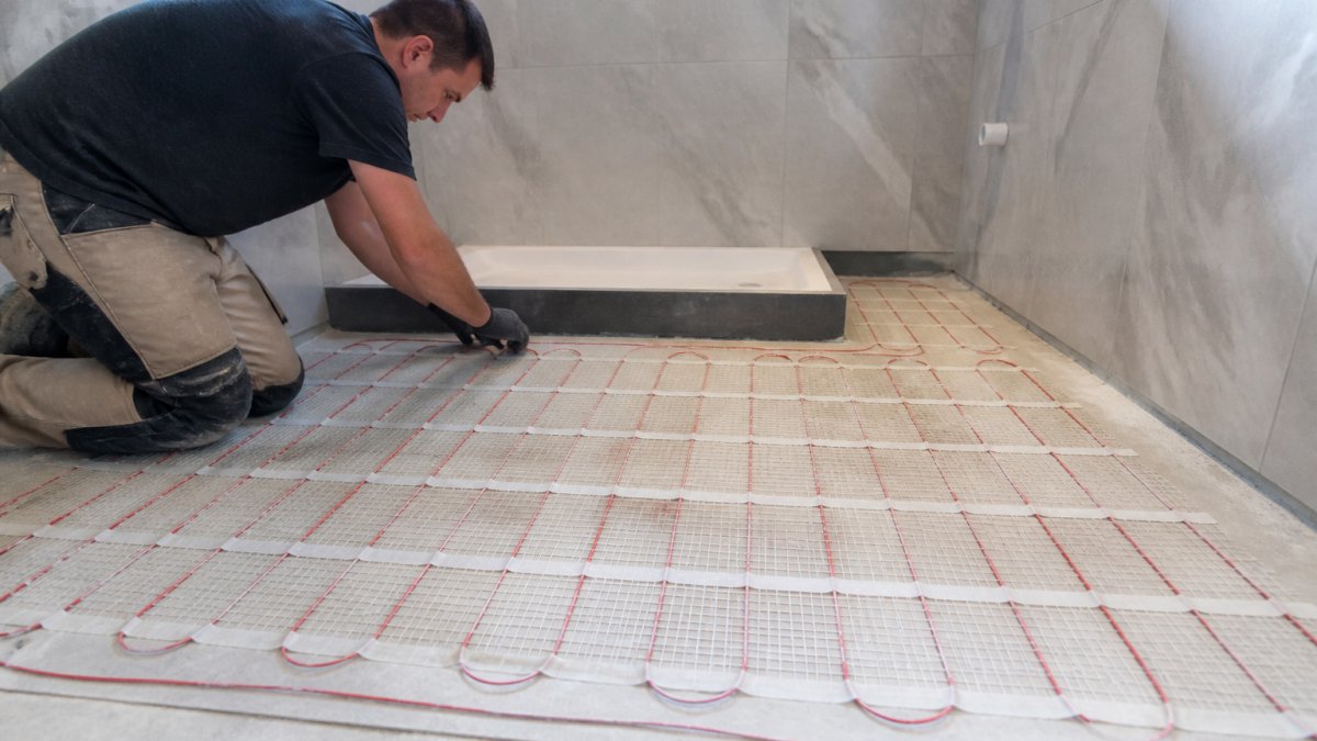 Professional tradesperson laying underfloor heating mat in bathroom during renovation, with heated cables visible on concrete subfloor before tile installation begins