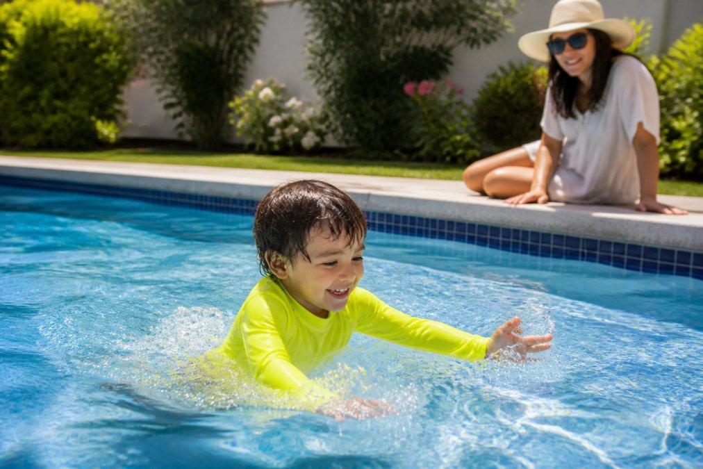 Young child wearing UPF 50 clothing playing in a sunny backyard pool while a parent watches from the edge