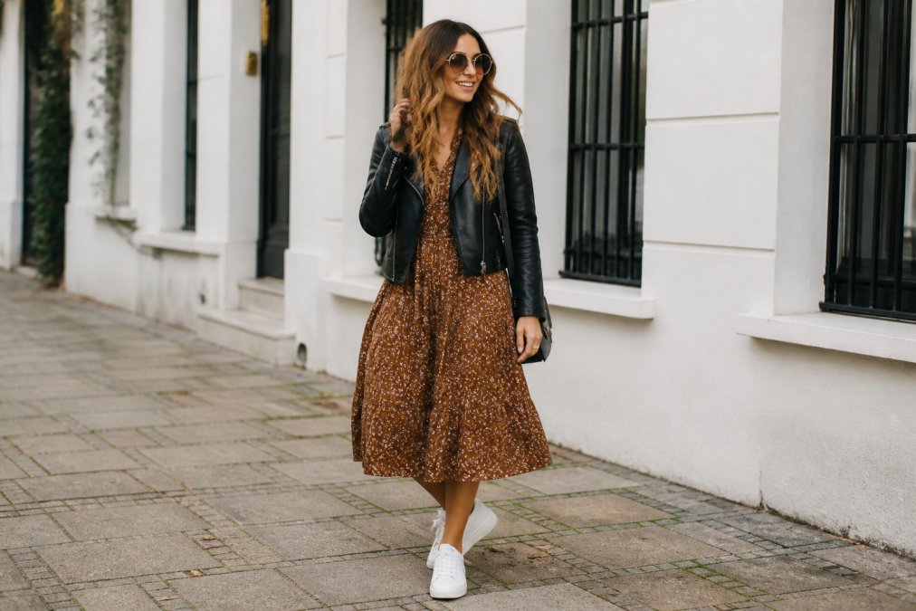 Person wearing a vintage-inspired 1970s-style dress in earth tones paired with contemporary white sneakers and a modern black leather jacket, styled for casual everyday wear in natural daylight