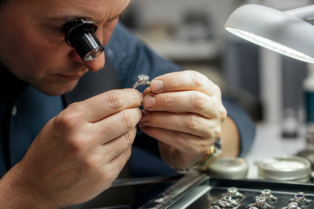 Professional jeweller's hands examining a vintage diamond ring under magnification with a loupe, studying hallmarks and gemstone quality under bright task lighting