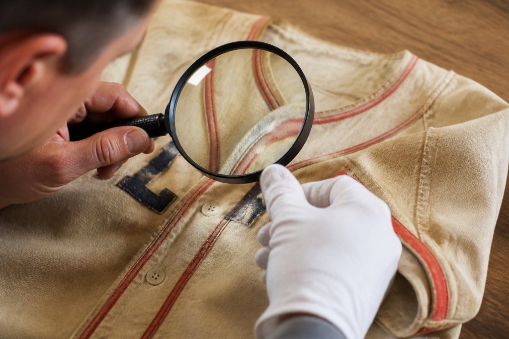 Close-up of a vintage sports memorabilia item being carefully examined with a magnifying glass under bright lighting, showing condition details, wear patterns, and historical markings on the fabric or surface