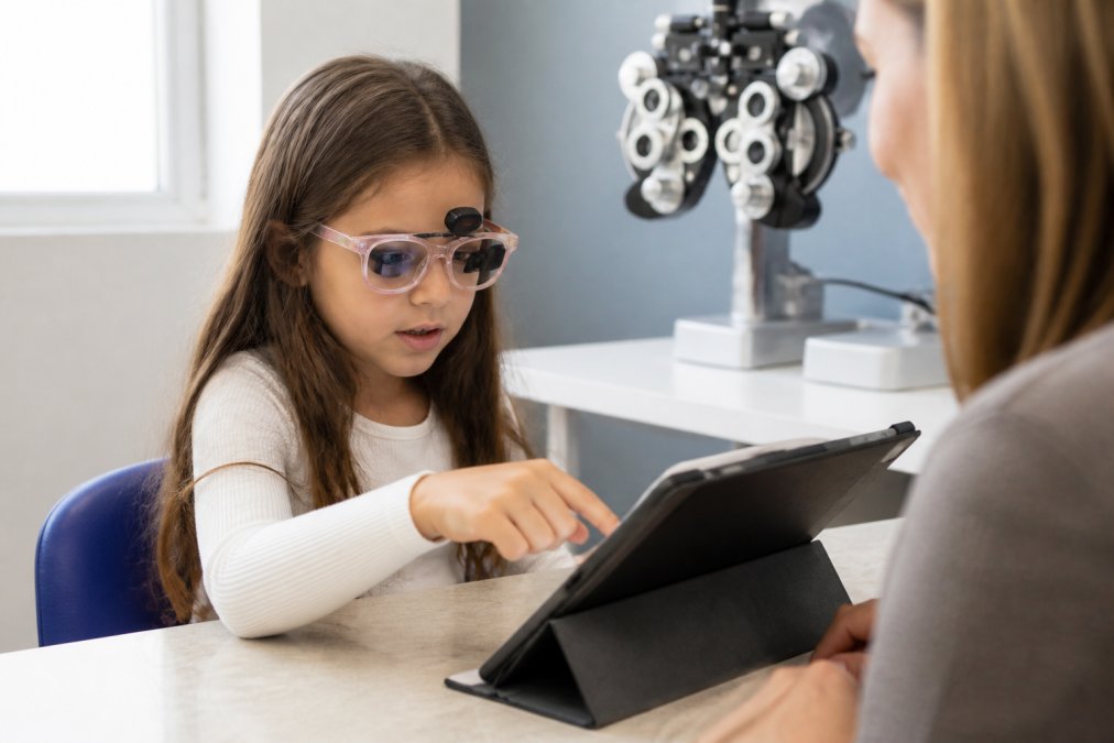 Child wearing specialized glasses during a vision therapy session, focused intently on a tablet displaying colorful therapeutic game with binocular targets, bright clinical lighting