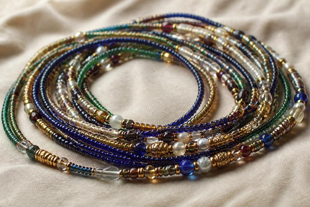 Close-up of intricate waist beads in rich jewel tones and metallic accents, displayed on a neutral background to showcase the detailed handwork and traditional color patterns