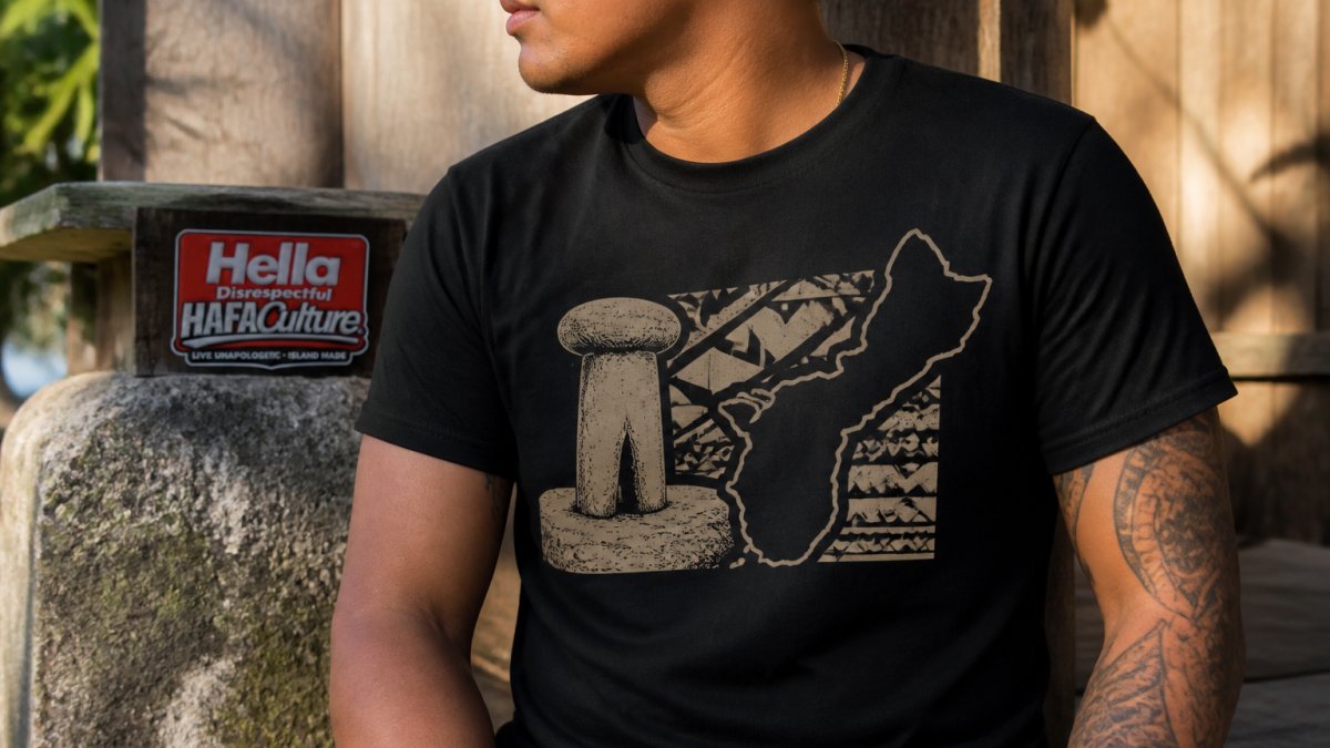 Close-up of a black t-shirt featuring a latte stone and island silhouette design, held by a person with Chamorro heritage, with warm natural lighting