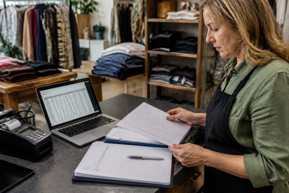 A retail store owner reviewing financial documents and a laptop showing cash flow spreadsheets at a checkout counter, with shelves of inventory visible in the background