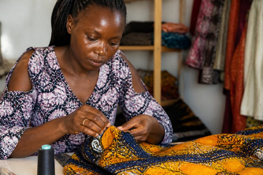 Female artisan handcrafting a garment with traditional textile techniques, hands working carefully with fabric and needle under natural studio lighting, showing meticulous attention to detail in ethical production