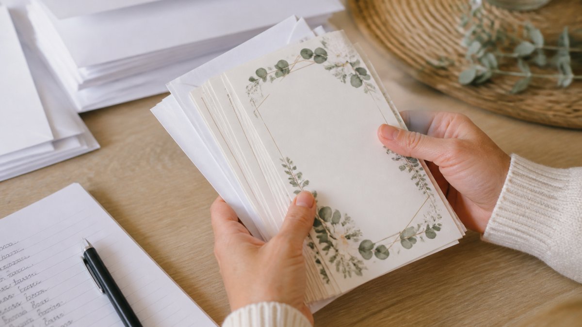 A close-up of hands holding a stack of freshly printed custom event invitations with white envelopes, arranged on a light wooden table with a pen and address list nearby