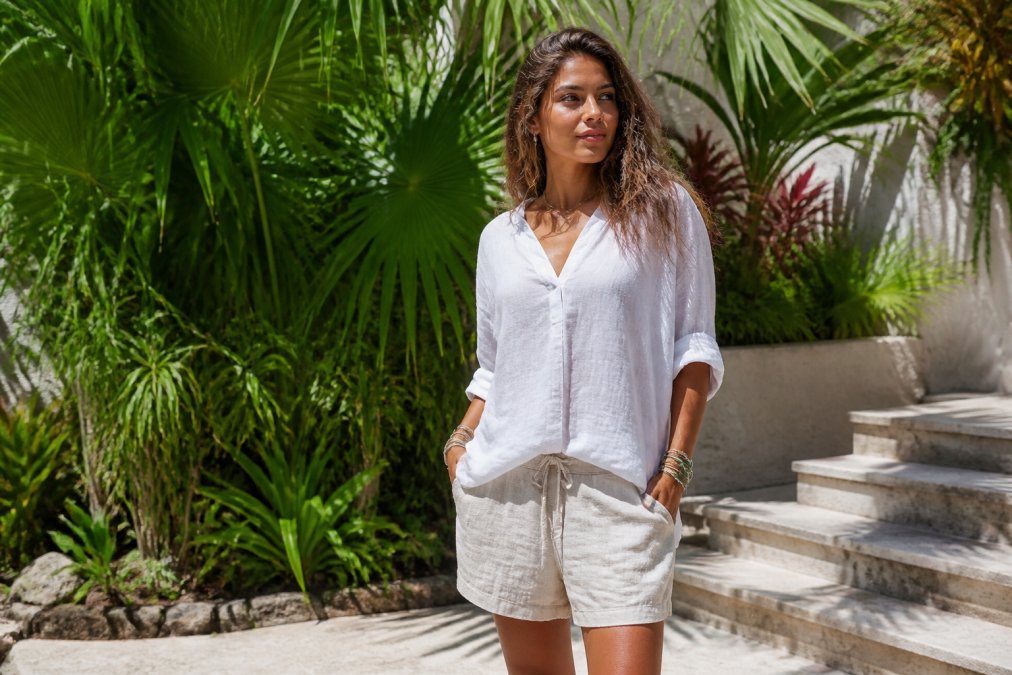 Woman wearing a lightweight linen tunic and linen shorts, standing in natural sunlight with tropical plants in the background, embodying effortless island aesthetic