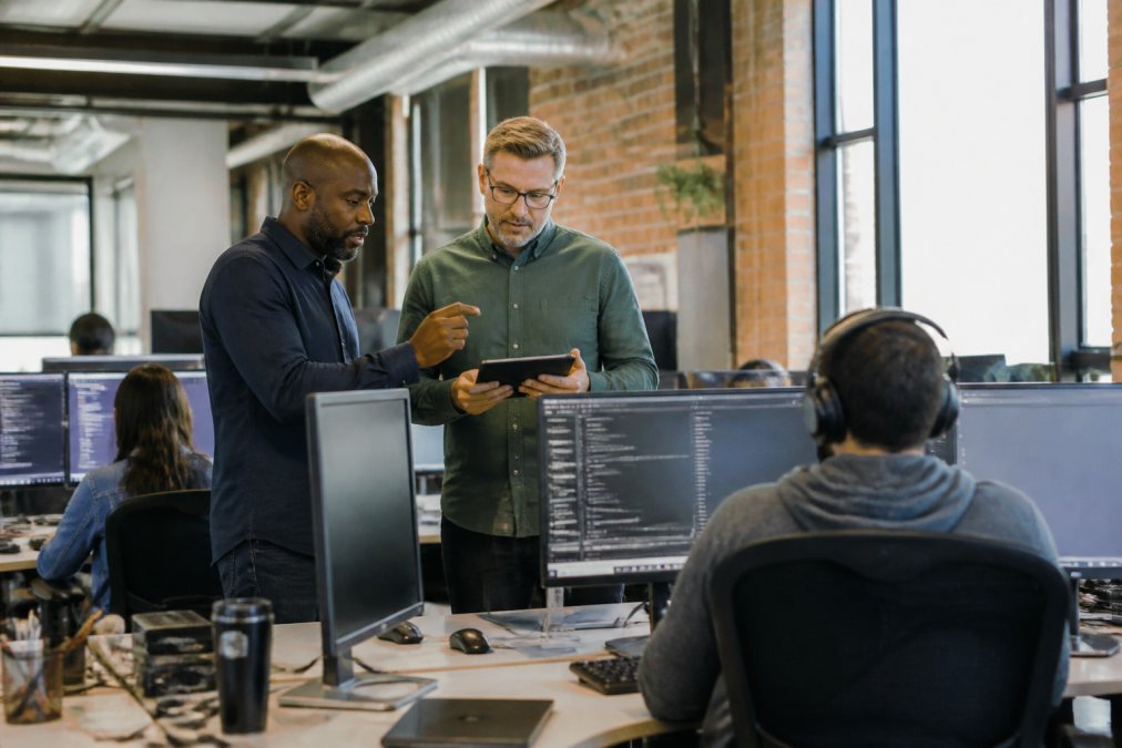 IT support team collaborating in a modern office environment with team members at computers reviewing performance metrics on a dashboard, manager discussing strategy with staff