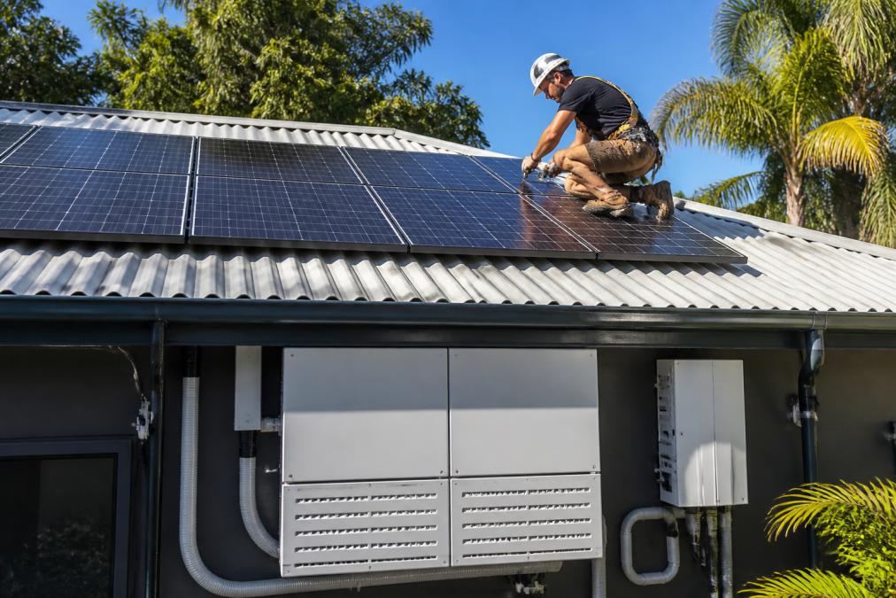 Licensed electrician installing solar panels on a residential rooftop in bright daylight, with battery storage equipment visible below, wearing safety harness and hard hat