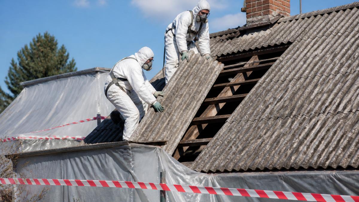 Dva pracovníci v bílých ochranných kombinézách a respirátorech demontují eternitové šablony ze šikmé střechy rodinného domu, kolem je položená ochranná fólie a výstražná páska, jasný den