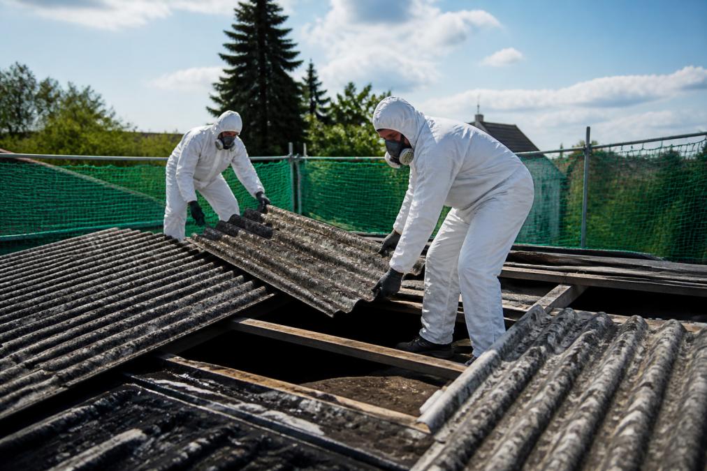 Certifikovaní pracovníci v bílých ochranných oděvech a respirátorech bezpečně demontují eternitové desky ze střechy pomocí speciálního vybavení, střecha je ohraničena bezpečnostními sítěmi