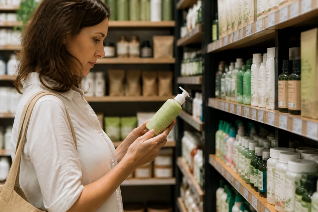 Žena v drogerii pečlivě otáčí produkt v ruce a čte drobný text na etiketě, kolem ní regály plné kosmetiky v přírodních barvách