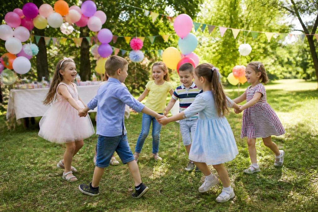 Grupa rozbawionych dzieci w wieku przedszkolnym bawi się w kole na świeżym powietrzu w słoneczny dzień, z kolorowymi balonami i dekoracjami w tle
