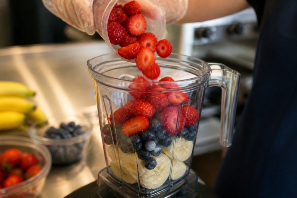 Close-up of fresh strawberries, blueberries, and banana slices being added to a commercial blender with visible fruit chunks and vibrant colors in natural kitchen lighting