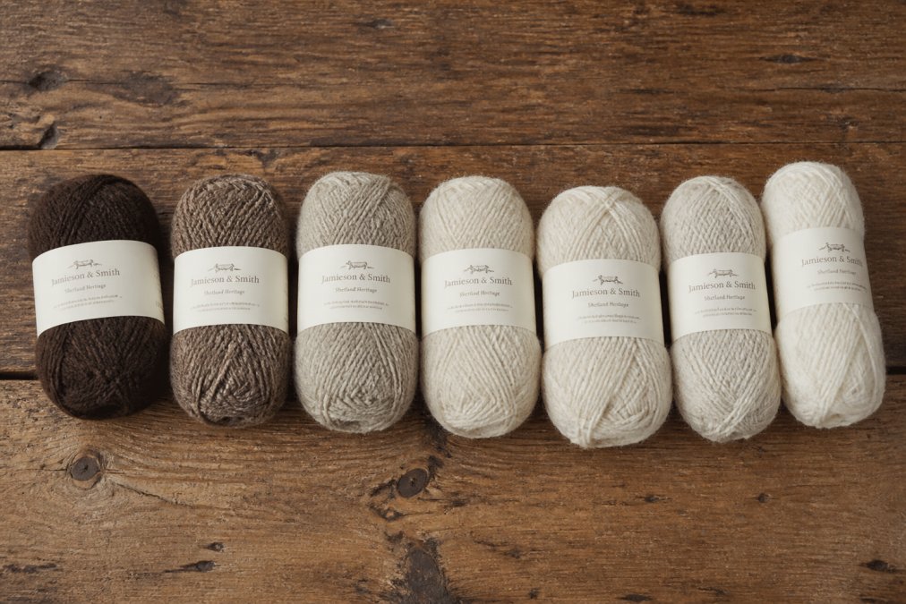 Flat lay of several skeins of natural-coloured Shetland wool yarn in varying weights arranged side by side on a worn wooden surface, with visible differences in ply thickness and fibre texture under soft natural light