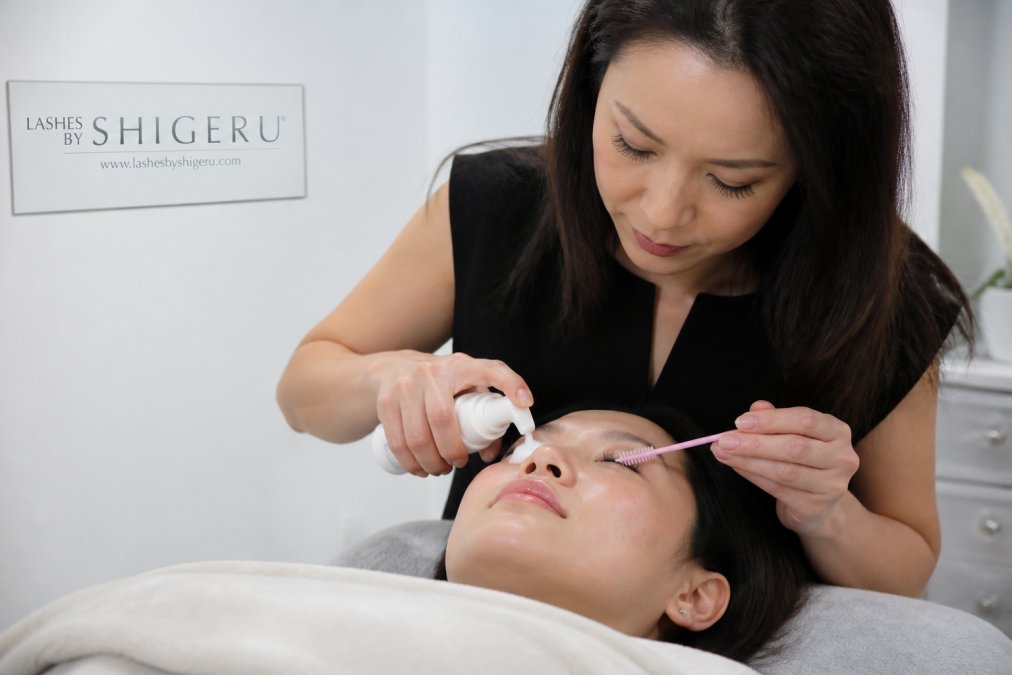 Hands gently cleaning lash extensions with an oil-free cleanser and spoolie brush, demonstrating proper maintenance technique in a bright, clinical setting