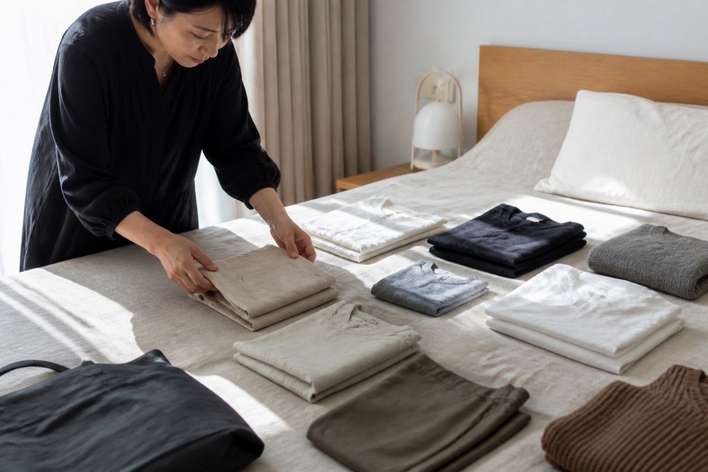 Woman arranging neutral linen and cotton clothes for a Japanese minimalist fashion capsule wardrobe on a bed