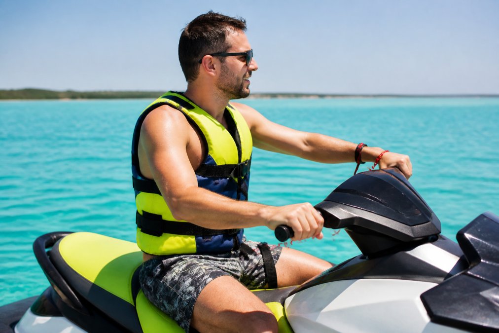 Jet ski rider wearing a bright-colored personal flotation device and safety lanyard, seated on a jet ski with calm turquoise water in the background, showing proper gear positioning and snug fit