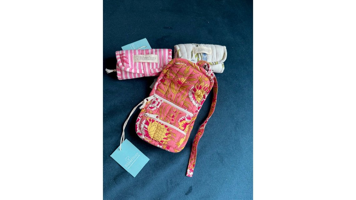 A person's hands carefully wrapping a small jewellery box in recycled tissue paper, with a cotton drawstring pouch and a sprig of dried flowers on a wooden table nearby