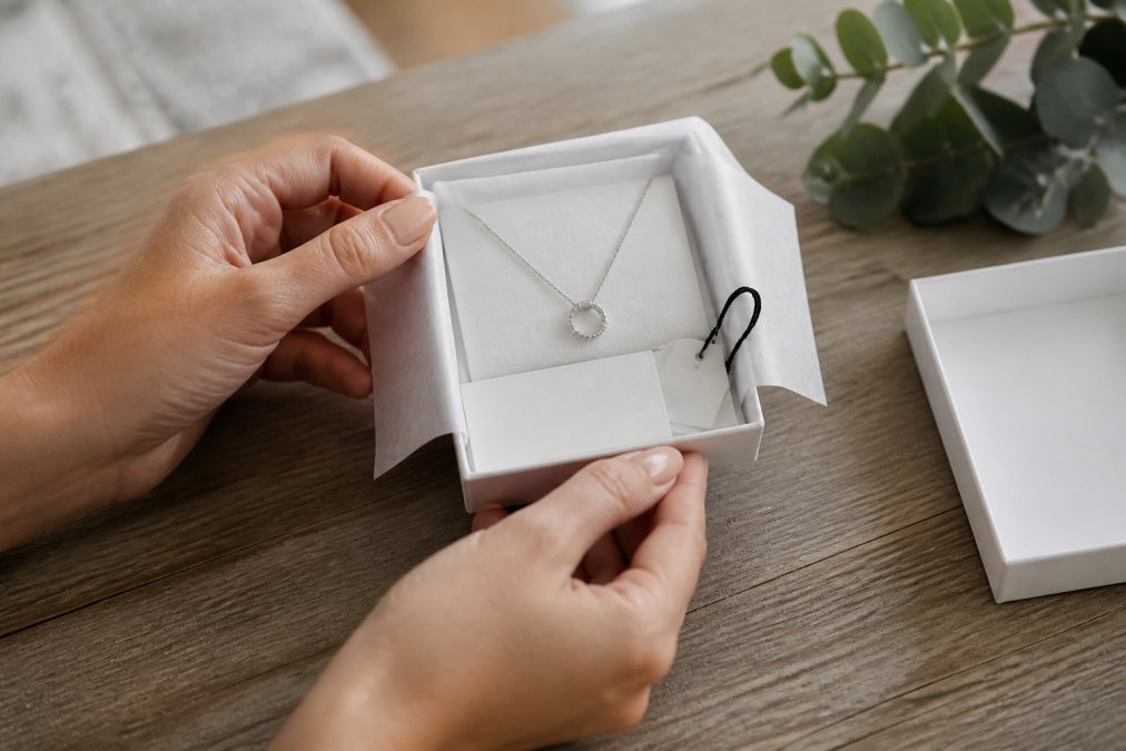 Close-up of hands carefully inspecting a delicate necklace still in its original packaging with protective tissue, care card, and branded tag visible under natural lighting