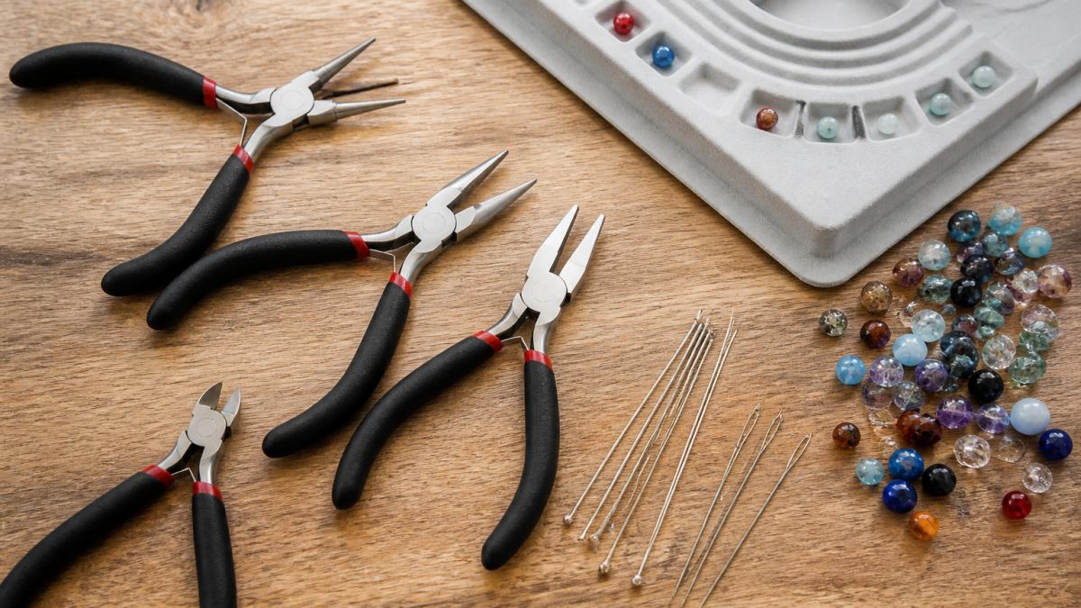 Flat lay of essential jewelry-making tools arranged on a wooden work surface: round-nose pliers, chain-nose pliers, flat-nose pliers, wire cutters, beading needles, and a bead mat with scattered colorful beads nearby