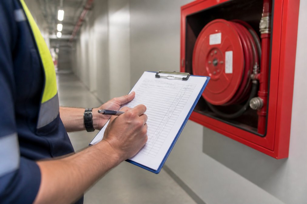 Close-up of a fire safety technician's hands writing detailed notes on a compliance checklist form attached to a clipboard, with a red fire hose reel cabinet visible in the background of a commercial building corridor under fluorescent lighting